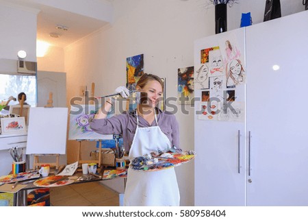 Cute female painter posing, laughing and fooling around in front of camera and smiling with brushes and colorful palette in hand, standing in studio with white art paintings on walls. Girl European