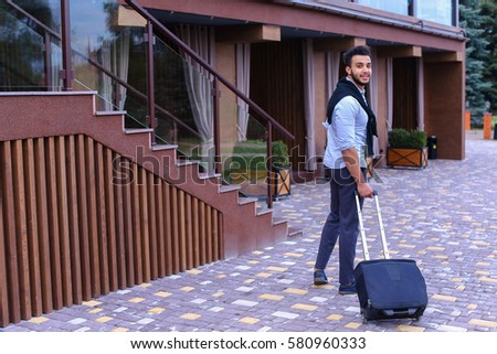 Arabic Muslim Young Man Coming Out of Corner and Goes With Travel Bag to Modern Restaurant, Stopped and Smiling, Holding Black Suitcase. Handsome Muscular Guy Dressed in Black Classic Trousers, White