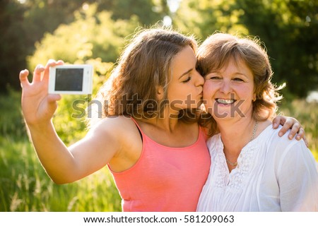 Adult daughter kissing her senior mother while taking selfie photo with mobile phone