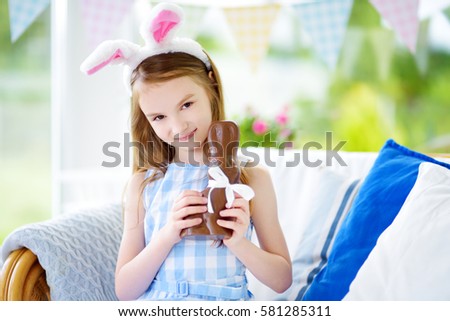 Cute little girl wearing bunny ears eating chocolate Easter rabbit. Kid playing egg hunt on Easter. Adorable child celebrate Easter at home. 