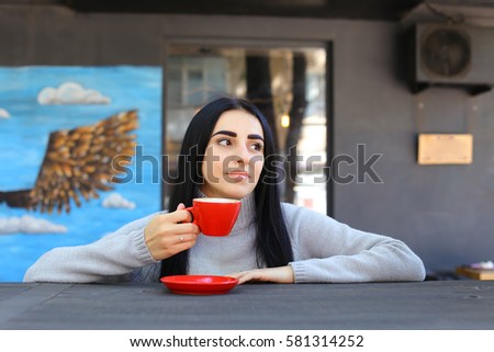 Charming young female girl smiling, holding red mug in hand, looks at camera, looks away, drinking coffee at wooden table on gray background panoramic windows and walls decorated with pictures. Girl