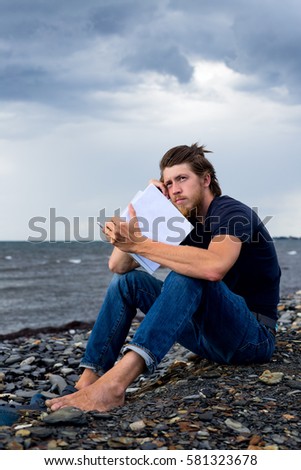 young handsome man holding new book and looking busy outdoors. Boy sitting on sea coast on grey day with heavy overcast over water