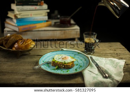 Black bread toast with fried egg, avocado and salmon spread in blue plate with coffee in background 