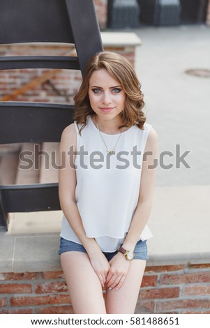Beautiful young girl dressed in a white blouse and denim shorts sitting on a brick wall