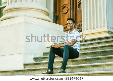 Way to Success. Wearing short sleeve striped shirt, black pants, leather shoes, African American Man sits on stairs of doorway on street in New York, works on laptop computer. Color filtered effect