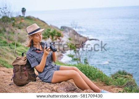 Photography and travel. Young woman in hat with rucksack holding camera sitting on cliff with sea view.