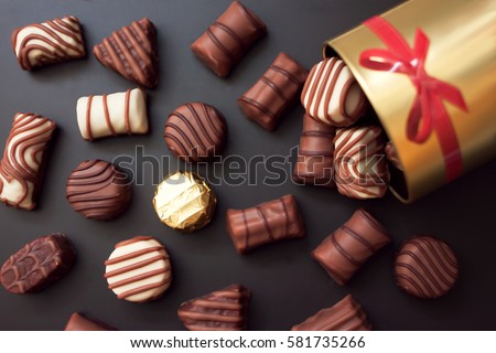 Chocolates in different shapes and colors spilled from the gold round box with a red bow on a black background. among all the sweets have a gold-wrapped in the color of the box. Royalty-Free Stock Photo #581735266