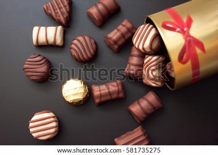 Chocolates in different shapes and colors spilled from the gold round box with a red bow on a black background. among all the sweets have a gold-wrapped in the color of the box Royalty-Free Stock Photo #581735275