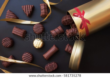 Chocolates in different shapes and colors spilled from the gold round box with a red bow on a black background. among all the sweets have a gold-wrapped. Golden ribbon between candies Royalty-Free Stock Photo #581735278