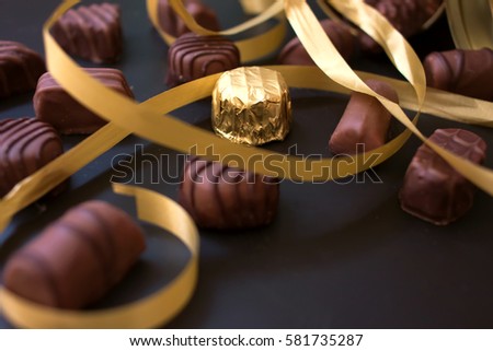 Chocolates in different shapes and colors spilled from the gold round box with a red bow on a black background. among all the sweets have a gold-wrapped. Golden ribbon between candies. Royalty-Free Stock Photo #581735287