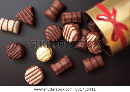 Chocolates in different shapes and colors spilled from the gold round box with a red bow on a black background. among all the sweets have a gold-wrapped in the color of the box Royalty-Free Stock Photo #581735311