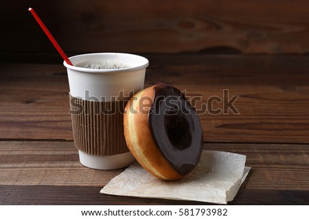 A chocolate frosted donut or doughnut leaning on a hot cup of fresh brewed coffee. 