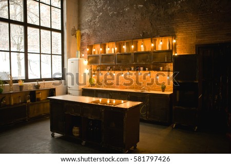 Large black metal-plastic window in the kitchen on the wall. Kitchen set, stove, sink, furnace.Spring or summer morning. The window shining bright sunlight. Dark loft interior