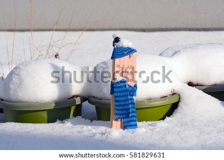 Wooden thermometer dressed up in winter clothes and placed in front of snow-covered plastic planters on terrace