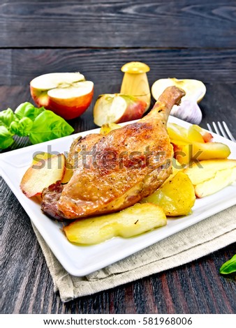 Roasted duck leg with apple, potatoes in a plate on a napkin, green basil, garlic and fork on a dark wooden board