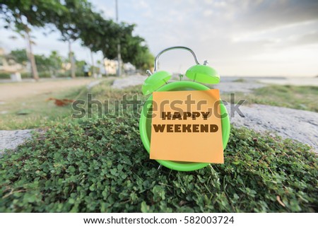 HAPPY WEEKEND word on a sticky note stick on analog clock over blurry beach and tree background