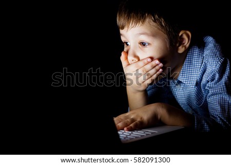 Young boy covered her mouth in fright, sitting at a computer on a dark night. The child is shocked by what he saw on the laptop. Watching a scary movie.