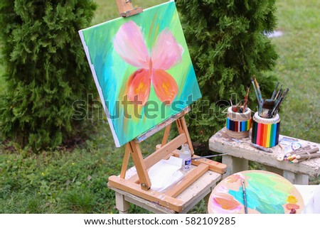 Photo completed and unfinished picture, works of art on which  image multi-colored butterfly on canvas and stands on wooden easel, palette and brush lying near easel, on table stands colorful banks i