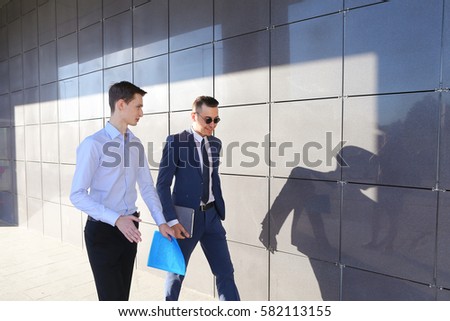 Two attractive confident young businessman male guys go and discuss important issues, hurry to a meeting, make decisions, solve problems, share business ideas, smiling, holding colored folders in