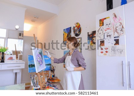 Artist attractive woman of European appearance writes oil painting, enjoying things what love , paint picture for sales and order, listening to music on headphones through phone, stands and works in