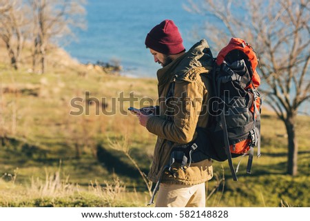 young handsome brutal bearded man traveling in the wild nature with touristic backpack, autumn style, warm coat, hat, traveler, adventure, free spirit, sea, holding looking at smartphone