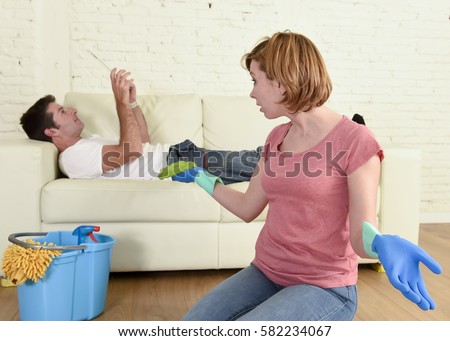 young couple with woman or wife kneeling washing and cleaning the floor scrubbing and man or husband lying on couch relaxed not giving her help in chauvinism and sexism concept Royalty-Free Stock Photo #582234067
