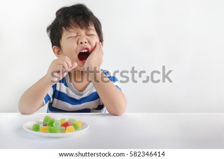 Sad little Asian boy suffering from toothache pain in mouth while eating candy, holding his cheek, dental pain. Royalty-Free Stock Photo #582346414