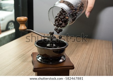 Pouring coffee beans into a rotary coffee grinder by the window and a glass of roasted coffee beans.