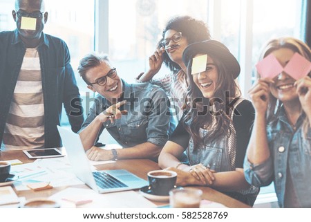 Group of beautiful young people at cafe table with laptop having fun together playing name game with sticky notes to their forehead, laughing Royalty-Free Stock Photo #582528769