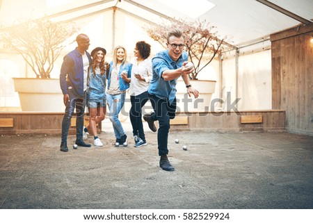 Young handsome man in glasses throwing steel petanque ball, his friends smiling standng behind him Royalty-Free Stock Photo #582529924