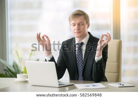 Young calm businessman working with laptop in yoga pose at modern office desk, minimize day discomfort, focusing on work, boost of energy after important task, keep feeling focused before meeting  Royalty-Free Stock Photo #582569848