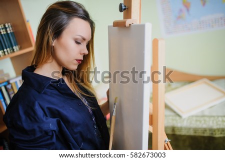 Attractive young woman artist begins to draw a new paint on a  wooden easel, holds brush.