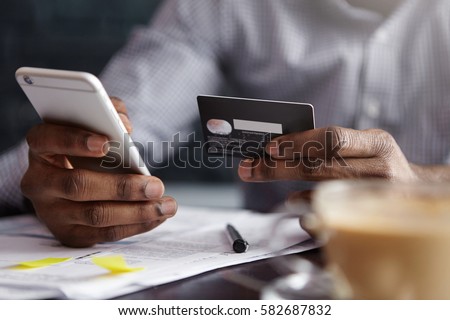 Cropped shot of African-American businessman paying with credit card online making orders via Internet. Successful black male holding plastic card making transaction using mobile banking application Royalty-Free Stock Photo #582687832