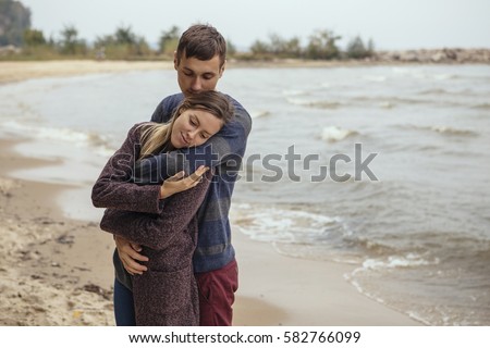 Happy thoughtful couple standing on a rock beach near sea hugging each other in cold foggy cloudy autumn weather. Copy space