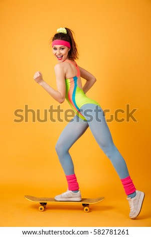 Full length of happy young sportswoman riding on skateboard over yellow background