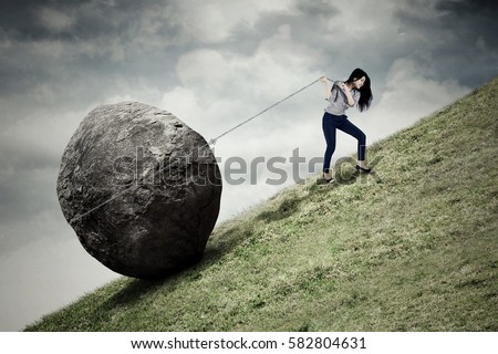 Image of young businesswoman climbing on the hill while pulling big stone with a chain Royalty-Free Stock Photo #582804631