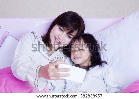 Cute little girl and her mother taking selfie photo together wit a mobile phone while lying on the bedroom