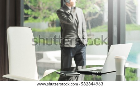 Laptop and tablet on glass table with businessman talking via mobile phone in background