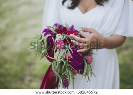 Happy bride in a simple white wedding dress holding a beautiful bouquet of flowers and green leaves. Woman in a stylish dress celebrating summer day wedding, blurred background, copy space.