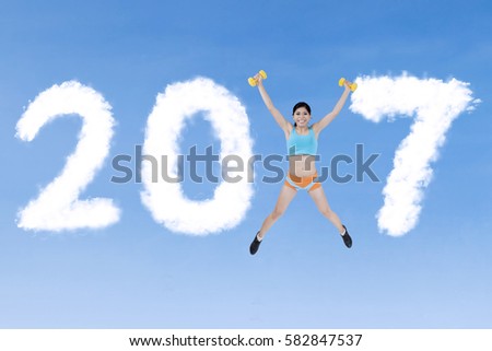 Healthy woman jumping on the sky while wearing sportswear and holding dumbbell with cloud shaped number 2017