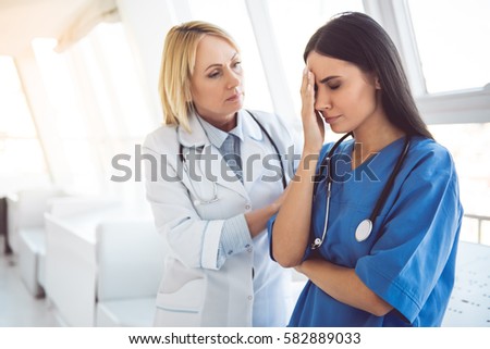 Beautiful doctor in blue scrubs is covering her face while her colleague is calming her Royalty-Free Stock Photo #582889033