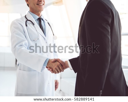 Cropped image of handsome mature doctor and young businessman shaking hands while standing in the hospital hall Royalty-Free Stock Photo #582889141