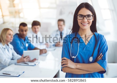 Beautiful young female medical doctor is looking at camera and smiling while her colleagues are sitting in the background