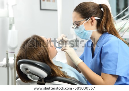 Dentist wearing eyeglasses gloves and mask examining a patient teeth with a dental probe and a mirror in a clinic box with equipment in the background Royalty-Free Stock Photo #583030357
