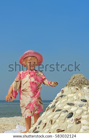 Beautiful little two-year girl standing on a sea beach near the sandy pyramid with shells on the background of a blue cloudless sky