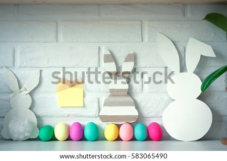 Three Easter white and retro bunny with colored eggs with sticky note against white brick wall background. Cute Easter celebration decoration. Copy space for text. Happy Easter card. Easter concept. 