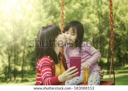 Young mother kissing her daughter on a swing while holding a smartphone and taking selfie picture
