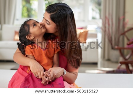 Pretty Indian woman kissing her little girl on cheek tenderly while spending free time together in living room