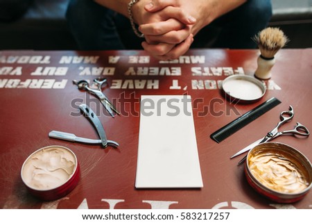 Blank card with barber tools on table free space. Side view on gules workplace with hairstyling instruments and hands of sitting man on background, Work, barbershop, manhood concept