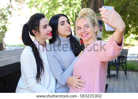Three wonderful young cute female girl girlfriends make selfie, photo on phone by heart, smiling and laughing in cafe on background trees outdoors. Girls with black hair dressed in white and gray